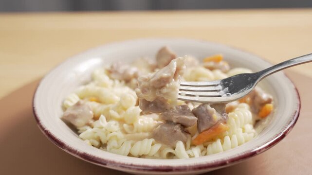 A hand with a fork picks up a piece of meat from a bowl of fusilli pasta with creamy stew and holds it in focus near the camera.