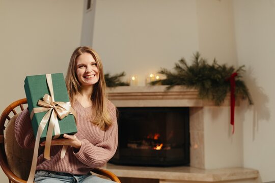 Happy woman holding christmas gift box by fireplace