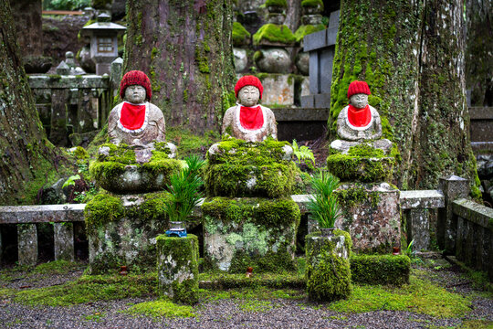 Three red-bibbed Jizo statues stand among moss-covered stones in a quiet Japanese forest cemetery