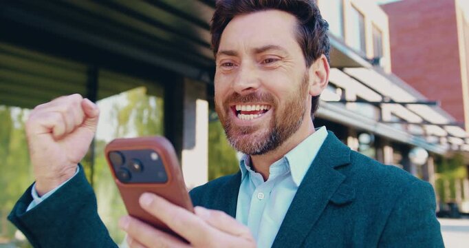 Close-up of a happy adult businessman outdoors looking at his smartphone screen, celebrating positive business news or a successful deal with enthusiastic hand gestures and joyful emotions.