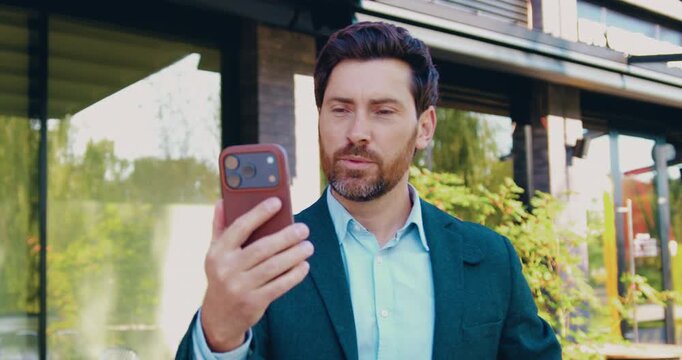 Adult businessman walking outdoors while making an online business call on smartphone