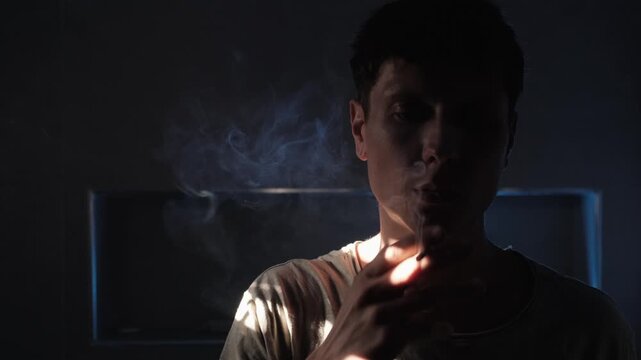 Addicted man smoking a cigarette alone in a dark room, surrounded by lingering smoke and harsh shadows, portraying nicotine dependence, compulsive habit and the difficult cycle of addiction