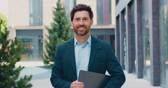 Smiling male businessman holding a laptop and posing outdoors while looking directly at the camera.