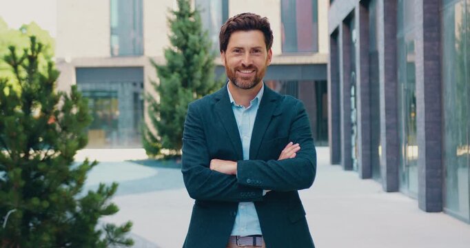 Adult bearded businessman standing outdoors in front of a modern office building, crossing his arms and smiling confidently while looking directly at the camera.