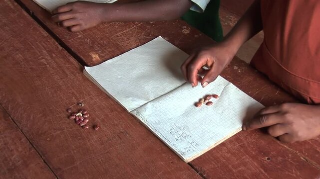 High-angle view of a child's hands arranging beans on a squared notebook at a wooden desk during a math lesson in a rural school in Kenya.