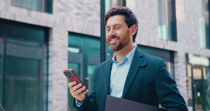 Businessman walking along a city street near a modern office building, holding a smartphone in his hand and looking to the side.