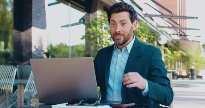Happy male professional working on a laptop while sitting at a table outdoors near an office building.