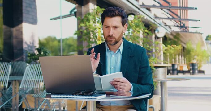 Professional man sitting at a table outdoors near an office, participating in an online video conference on his laptop while taking notes in a notebook.