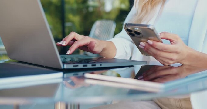 Close-up of a female hands using a smartphone and typing on a laptop while working remotely outdoors, transferring information between devices and managing digital tasks.