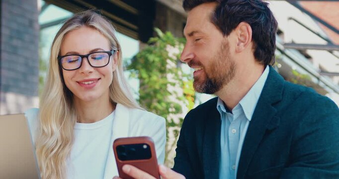 Smiling business colleagues working on laptop and sharing smartphone Information outdoors