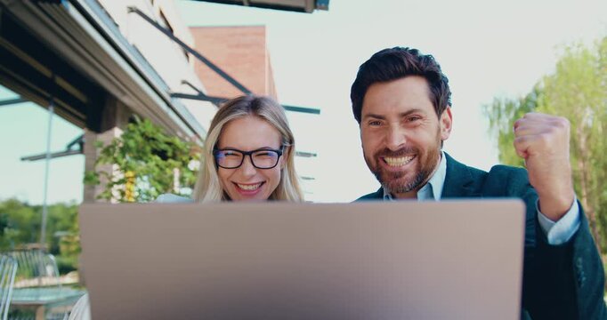 Smiling businessman and businesswoman celebrating positive results while looking at a laptop screen during an outdoor meeting near an office building