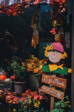 wooden scarecrow welcome sign among orange autumn leaves, rustic fall harvest decoration featuring hanging corn and floral storefront display