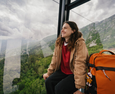 Smiling young woman sitting on bench inside cable car cabin next to her backpack