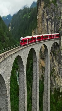 Red train crossing a high stone viaduct bridge through the swiss mountains