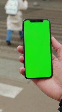Person holding a smartphone with a blank green screen outdoors close up view