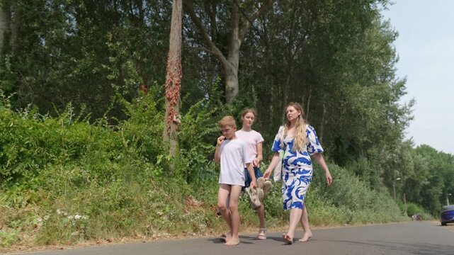 Woman and two children are walking barefoot along a tree lined road slow motion family walk the boy is biting his fingers