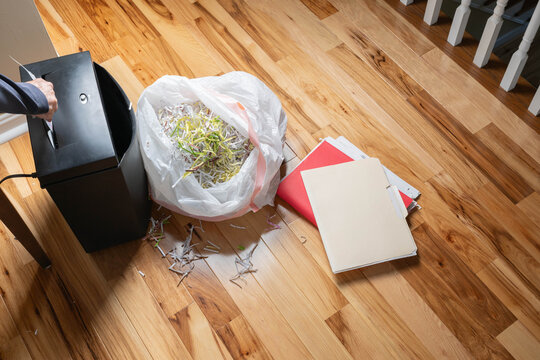Shredding paper documents, destroying sensitive, personal data.  Shredder, stack of file folders, and bag of shredded paper on hardwood floor.