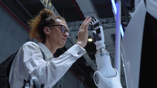 Young man wearing glasses gives a high five to a humanoid robot, demonstrating human robot interaction and advances in robotics.