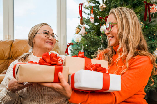 Two women exchange wrapped gifts beside a Christmas tree
