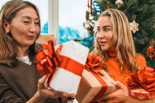 Two women exchange wrapped gifts beside a Christmas tree