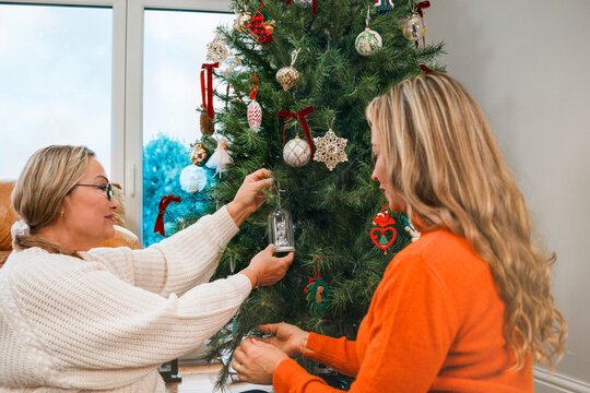 Two women decorate a Christmas tree beside a window