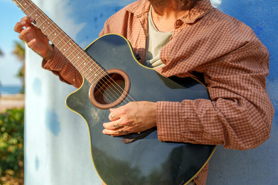 Musician playing a dark acoustic guitar outdoors, tattooed hands strumming strings in warm sunlight beside a blue wall