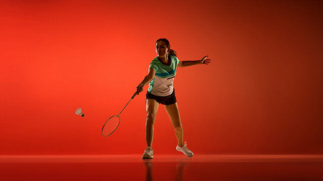 Focused woman making low forehand badminton shot in orange studio. Beginner sports lessons, activewear shopping, leisure centers concept.
