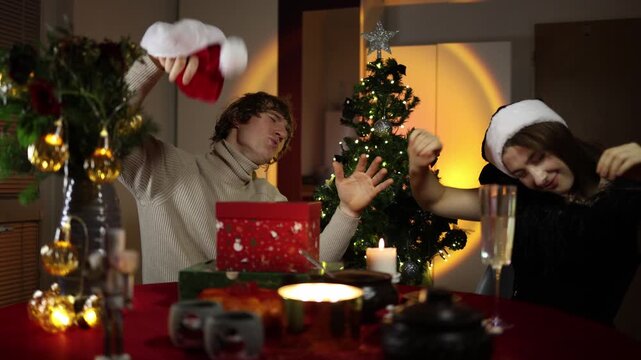 A young couple dances and lets loose at a Christmas and New Year party at home as the man waves a Santa hat and the woman sways happily beside a glowing holiday tree late