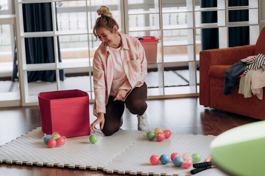 Young woman kneeling beside storage ottoman amid bright play balls shows realistic household reset and low pressure cleanup in comfortable urban apartment setting across modern household culture.