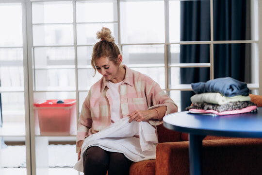 Woman holding pale textile next to neatly stacked garments in living room conveys attentive fabric inspection and care decisions that extend everyday clothing usability while highlighting repeat wear.