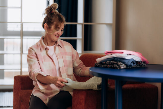 Woman holding soft green knit garment above stacked clothes on blue side table demonstrates practical seasonal wardrobe rotation and low waste domestic fashion habits across modern household culture.
