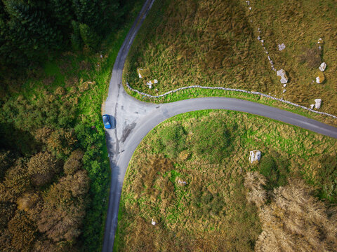 Aerial view shows winding rural road splitting into two paths. Blue car parked on intersection surrounded by greenery. Lush grass and trees frame the scene. Stone wall lines one side