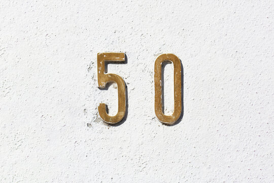 Number 50 vintage brass house number on textured plaster wall in strong sunlight, worn metal digits with scratches, address sign detail for real estate, architecture and street numbering concepts