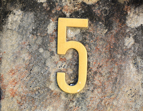 Number 5 golden brass house number mounted on weathered stone wall with rusty stains and lichen, aged metal digit for home address signage, real estate, architecture and street numbering concepts