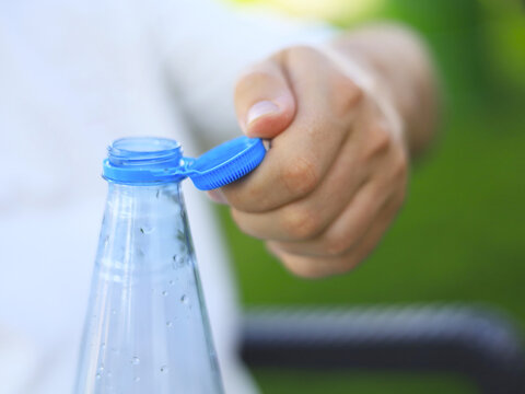 Hand holds a blue tethered cap beside an open water bottle, with beverage packaging designed to keep its plastic closure attached to the container as the consumer opens a drink for hydration outdoors.
