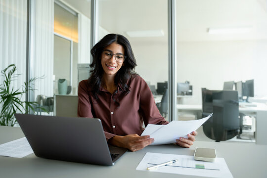 Smiling young woman analyst reviewing report and documents at office desk with laptop and phone, Indian, Middle Eastern or multiracial business financier and economist in 30s comparing chart data