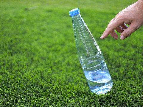 Hand reaching toward a capped water bottle on grass, with clear plastic beverage packaging that keeps drinking water portable for outdoor hydration during breaks from exercise and recreation.