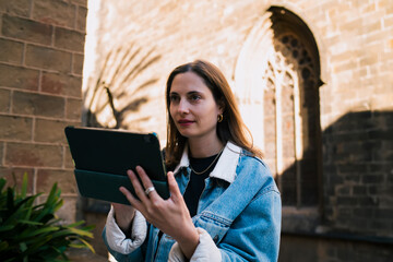 Focused Caucasian female student holding digital tablet computer outdoors near historic architecture, engaging with online education distance learning platforms and e-learning apps © BullRun