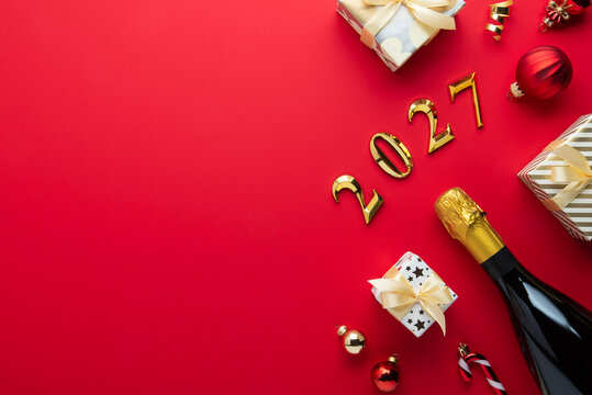 Merry Christmas and Happy Holidays greeting card, frame, banner. New Year 2027. Red and golden Christmas ornaments and gifts, presents with champagne on red background top view. Copy space.