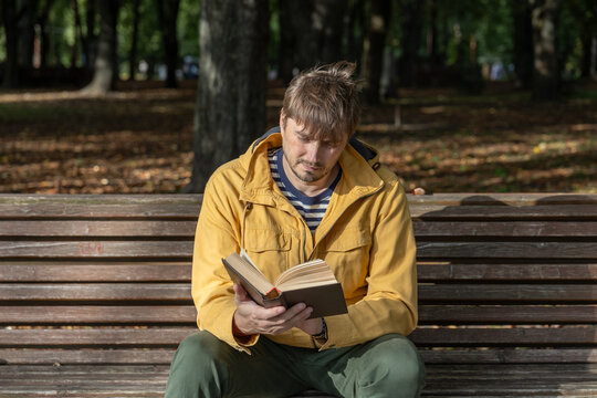 Man reading book while sitting on wooden bench in park, enjoying literature and outdoor reading