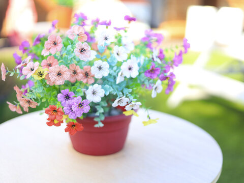 Colorful artificial flowers in a red pot sit on a white table as a floral centerpiece, combining tabletop home decor with a compact arrangement suited to small spaces and casual seating area styling.