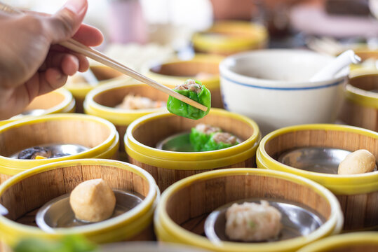 Close up of hand holding chopsticks picking green pork dumpling over traditional dim sum bamboo steamers