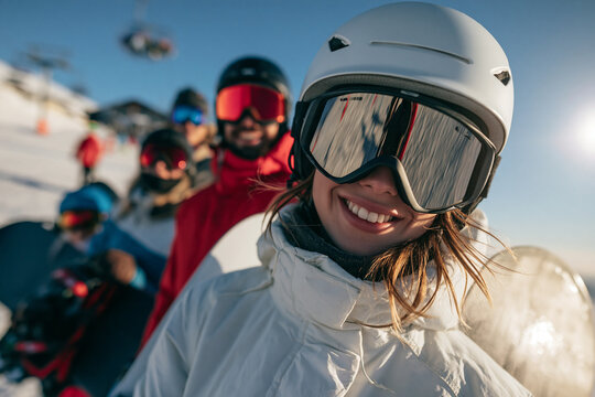 A smiling woman wearing a white helmet and goggles stands in the foreground with friends lined up behind her on a sunny snowy mountain slope ready for a day of snowboarding adventure