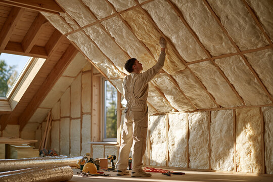 A focused worker in beige overalls installs fiberglass insulation batts between the wooden rafters of an attic bathed in warm natural sunlight streaming through a window during construction