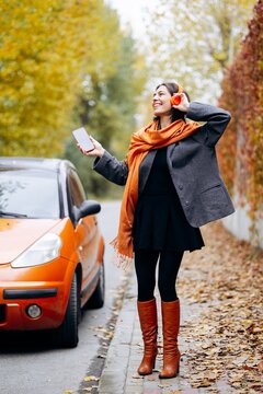 Woman enjoying music on an autumn street, holding a smartphone and adjusting her headphones beside an orange car