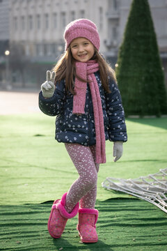 Happy Young Girl Wearing Pink Winter Hat, Scarf and Boots Smiling and Making Peace Sign Outdoors in a Winter Park