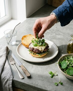 Chef Hand Placing Microgreens On Juicy Cheeseburger At Kitchen Counter