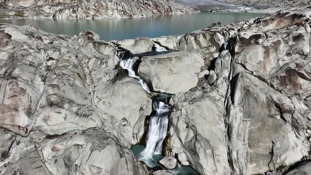 Dynamic aerial drone footage capturing the powerful waterfall of glacial meltwater cascading down steep alpine rocks from the Rhone Glacier lake near Furka Pass, Switzerland. Scenic drone shot showcas