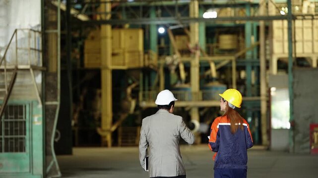 Wide rear shot of the rice mill manager and a female engineer walking deeper into the hall toward towering yellow milling machinery, he points up with a pen while she looks