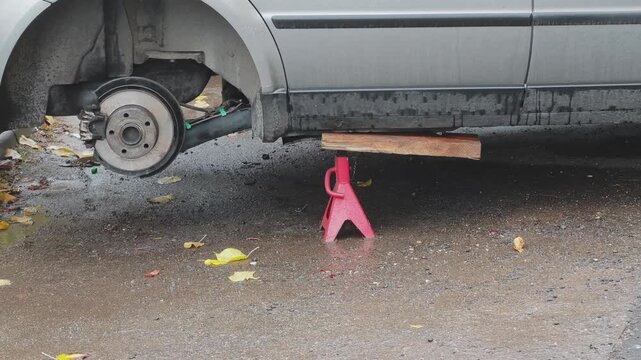 Passenger vehicle resting on jack stand with removed rear wheel, exposing brake rotor assembly on wet asphalt surface